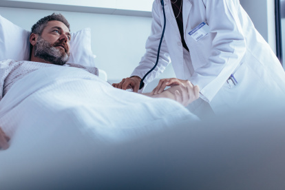 Hospitalized man lying in bed while doctor checking his pulse. Female physician examining male patient in hospital room.