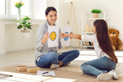 Professional children"s psychologist talking to little kid about emotions. Girl chooses happy smiley emoticon from two EQ cards that female therapist shows her during interview appointment meeting