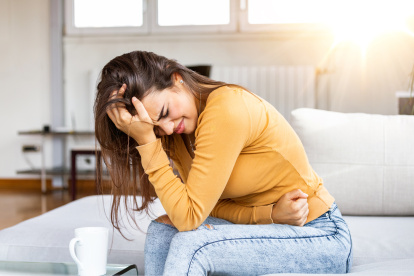 Young sick woman with hands holding pressing her crotch lower abdomen. Medical or gynecological problems, healthcare concept. Young woman suffering from abdominal pain while sitting on sofa at home