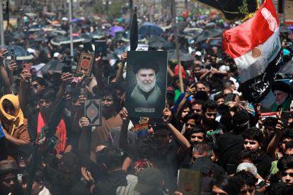 Los musulmanes chiítas iraquíes cantan eslóganes y llevan una imagen de Muqtada al-Sadr y copias del Corán mientras el humo está en el aire durante una reunión después de las oraciones del viernes en la ciudad de Sadr en Bagdad, Irak.