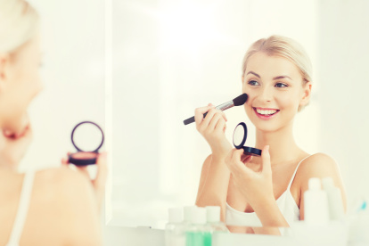 beauty, make up, cosmetics, morning and people concept - smiling young woman applying blush with makeup brush and looking to mirror at home bathroom
