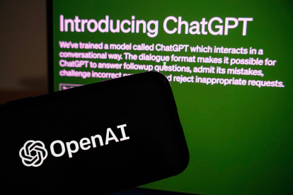 Vista de la página de ChatGPT detrás de un teléfono móvil en el que se ve el logo de la empresa OpenAI