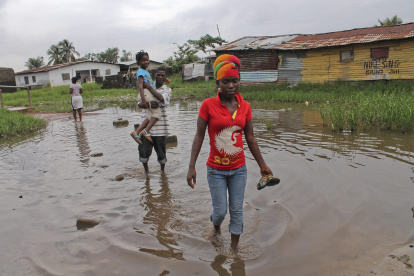 Un grupo de vecinos, con los pantalones remangados y el calzado en la mano, cruzan una zona encharcada.