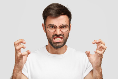 Portrait of emotive annoyed irritated unshaven male clenches teeth and gestures angrily while argues with wife, has irritated negative facial expression, wears casual white t shirt, stands indoor