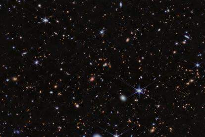 Imagen del protocúmulo Telaraña visto por la NIRCam (Near-InfraRed Camera) de Webb. Cientos de galaxias aparecen en esta vista, que se sitúa sobre el fondo negro del espacio. Hay muchos objetos superpuestos a distintas distancias. Incluyen grandes estrellas azules en primer plano, algunas con ocho picos de difracción, y galaxias espirales y elípticas blancas y rosas. En toda la escena aparecen numerosos puntos diminutos de color naranja]. Crédito: