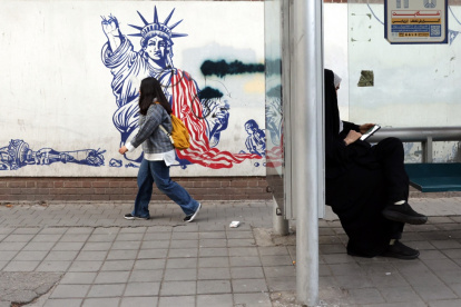 Una mujer iraní con velo se sienta en una estación de autobuses junto a un mural antiestadounidense cerca de la antigua embajada de Estados Unidos en una calle de Teherán.