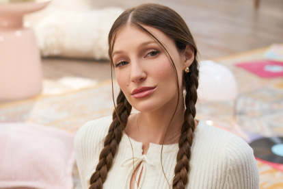 Fotografía promocional de Yami Safdie en un ambiente interior con tonos pastel. La artista aparece sentada en el piso, con trenzas, blusa blanca tejida y una estética visual vinculada al universo de Querida Yo.