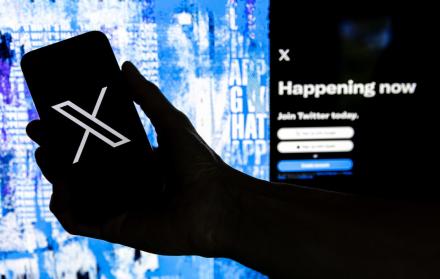 Fotografía que muestra un usuario mientras sostiene un teléfono móvil que muestra el logotipo 'X' frente a la página principal de Twitter.