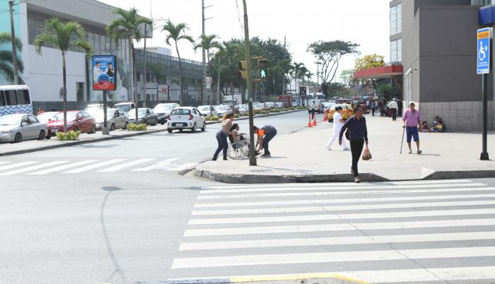 Rampas. Una mujer en silla de rueda es ayudada a subir la acera sin rampa. Al frente, una subida demasiado estrecha.