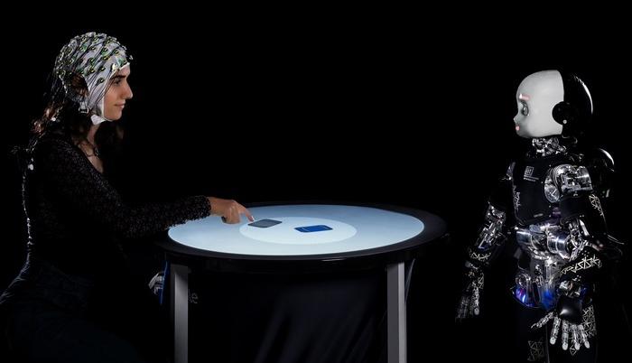 Una persona y un robot humanoide participan en un juego competitivo, mientras se mide su actividad cerebral con un electroencefalograma.
