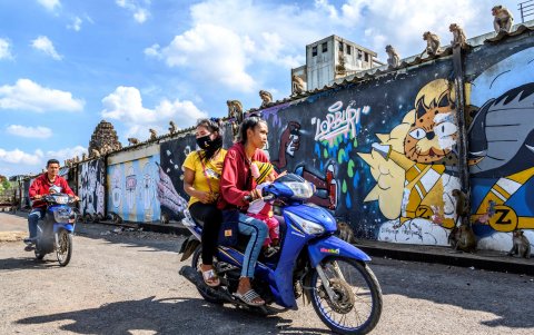 LOPBURI. Una pareja se traslada en motocicleta por una calle de esta ciudad, mientras los monos toman sol en el filo de una pared.