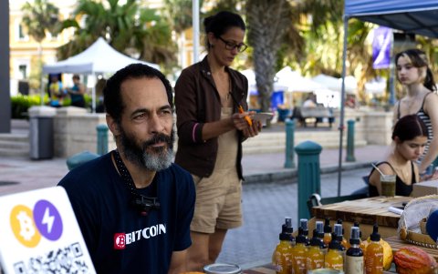 José Crespo posando junto a carteles de aceptación de bitcoin como forma de pago, en el Mercado Agrícola del Viejo San Juan (Puerto Rico).