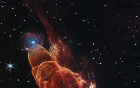 La nube de forma cónica de color rojo anaranjado corresponde a Herbig-Haro 40/50, en el extremo superior izquierdo hay una galaxia de fondo en forma de espiral con un centro azul.