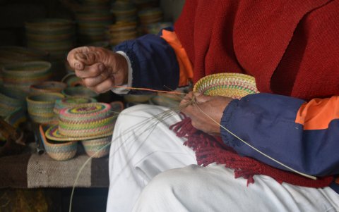 José teje con paciencia en la entrada de su hogar en Cuatro Esquinas, rodeado de sus creaciones de paja blanca.