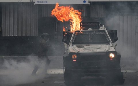 Un policía antidisturbios se encuentra junto a un vehículo incendiado durante enfrentamientos con manifestantes.