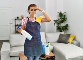 Young brunette woman wearing apron holding cleaning products at home worried and stressed about a problem with hand on forehead, nervous and anxious for crisis