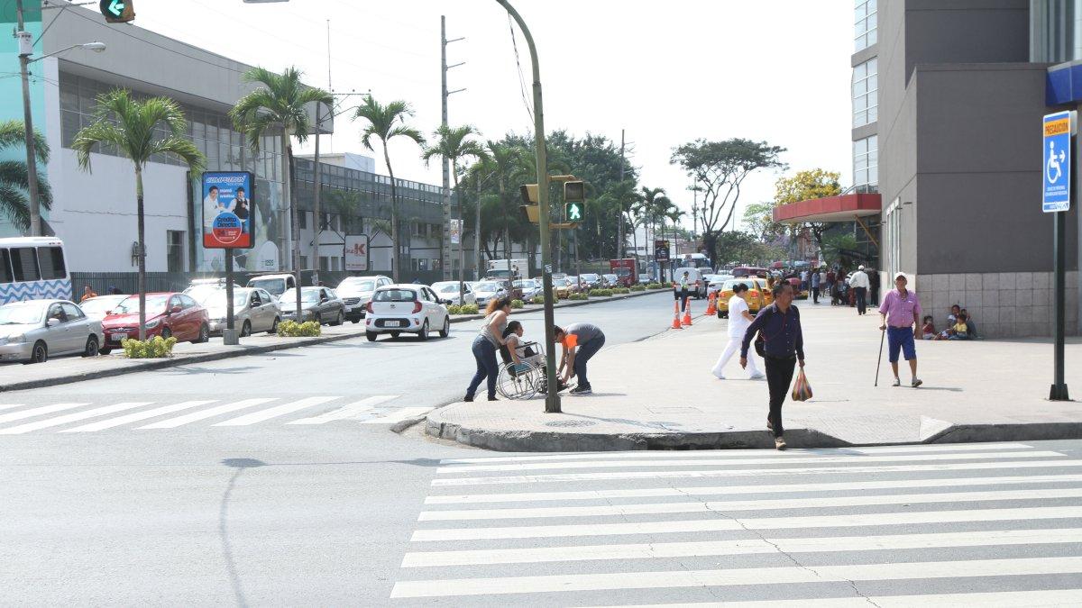 Rampas. Una mujer en silla de rueda es ayudada a subir la acera sin rampa. Al frente, una subida demasiado estrecha.