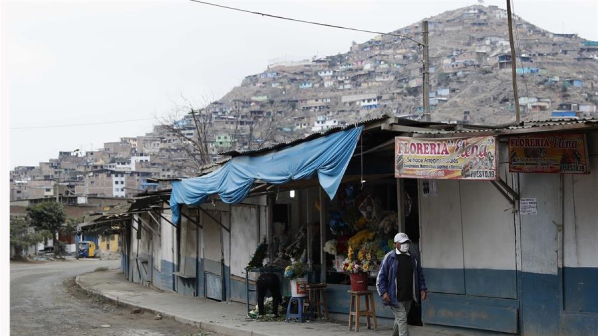 Un hombre con mascarilla camina ante un puesto de venta de arreglos florales en las inmediaciones del Cementerio de Nueva Esperanza del distrito de Villa María del Triunfo, en Lima, capital de Perú.