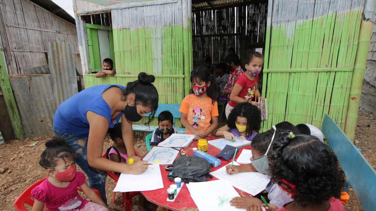 En una casa de caña, con pisos de tierra, 20 niños reciben ayuda educativa en la cooperativa Realidad de Dios. Líderes comunitarios colaboran en la tarea.