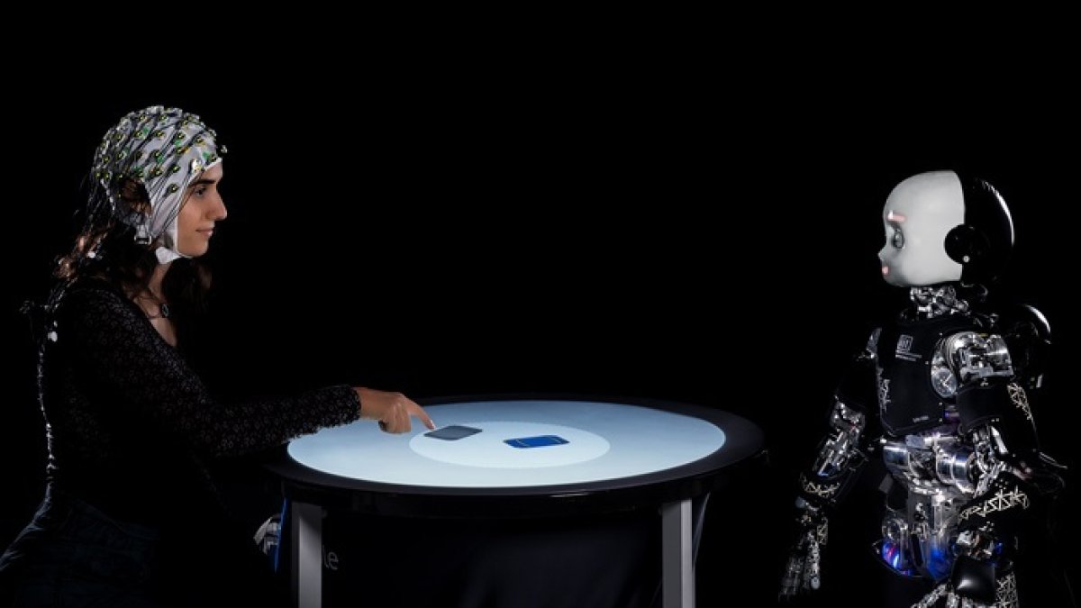 Una persona y un robot humanoide participan en un juego competitivo, mientras se mide su actividad cerebral con un electroencefalograma.