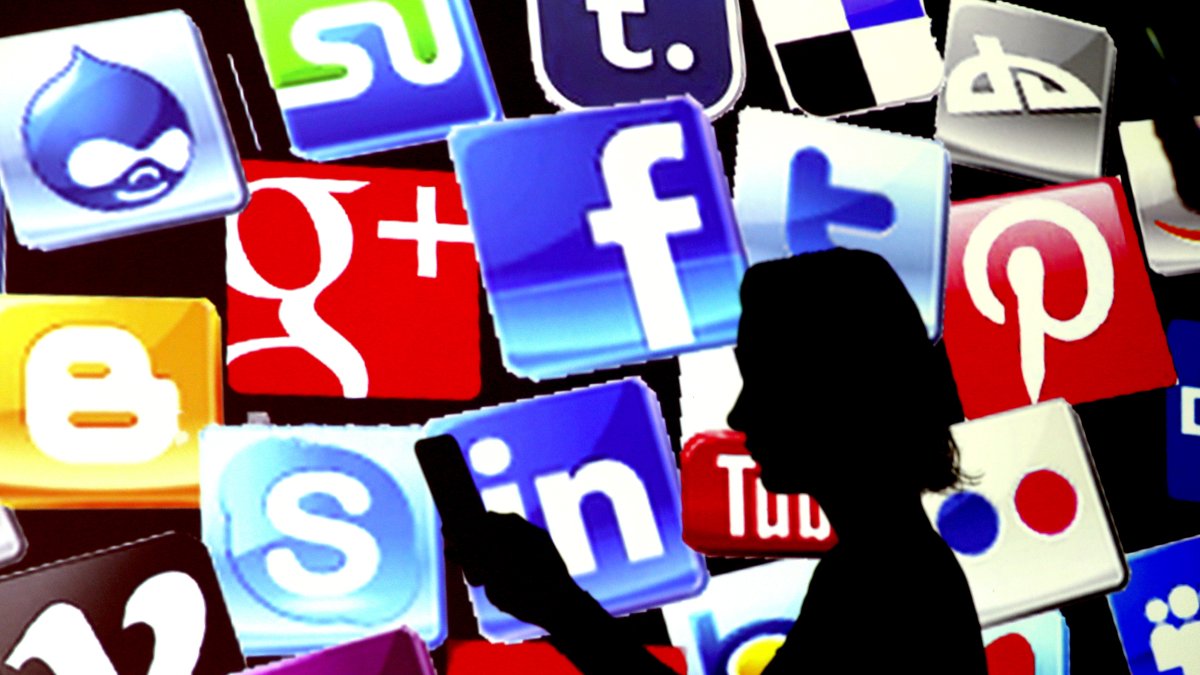 Una mujer revisa su teléfono frente a una pantalla que muestra los íconos de las redes sociales más populares.