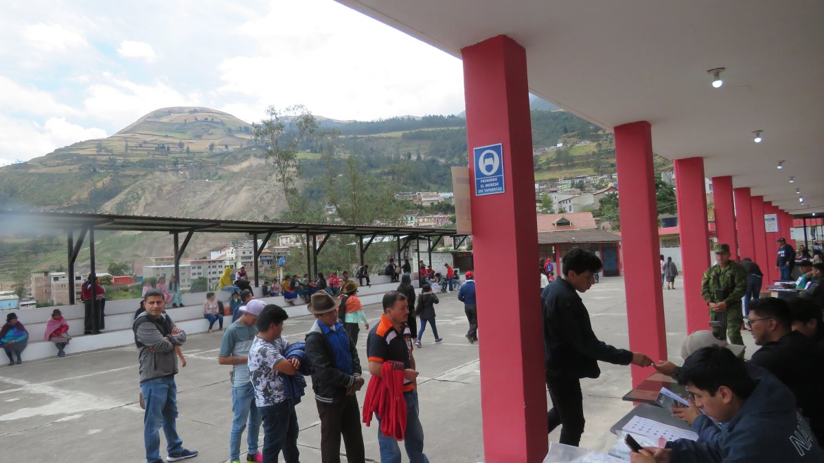 Alausí. Varias personas hacen fila en un recinto electoral. Al fondo, se observa la zona cero del derrumbe.