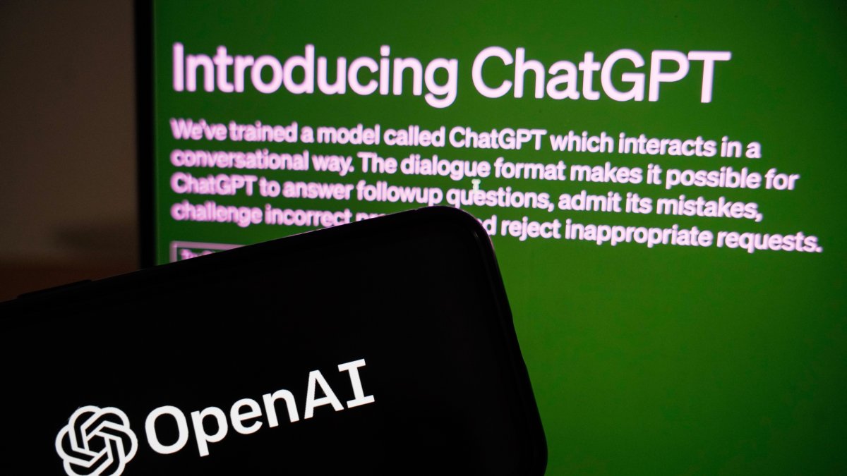 Vista de la página de ChatGPT detrás de un teléfono móvil en el que se ve el logo de la empresa OpenAI