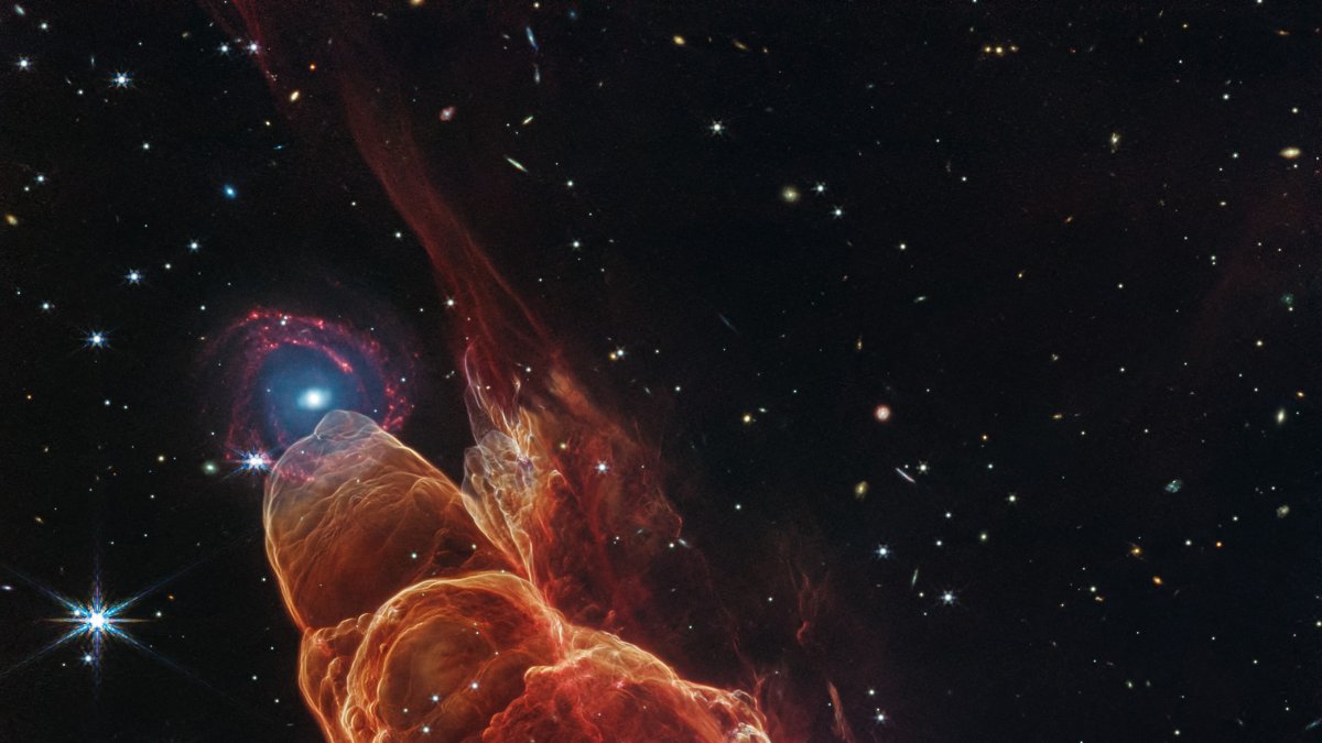 el telescopio espacial James Webb. La nube de forma cónica de color rojo anaranjado corresponde a Herbig-Haro 40/50, en el extremo superior izquierdo hay una galaxia.