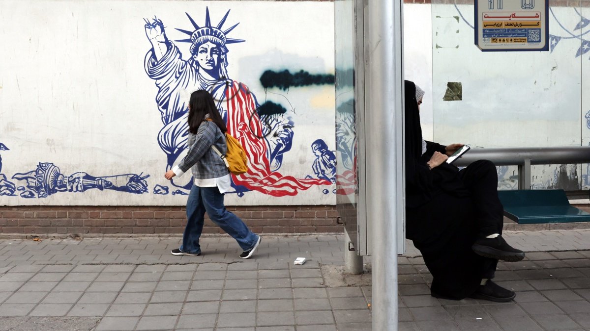 Una mujer iraní con velo se sienta en una estación de autobuses junto a un mural antiestadounidense cerca de la antigua embajada de Estados Unidos en una calle de Teherán.