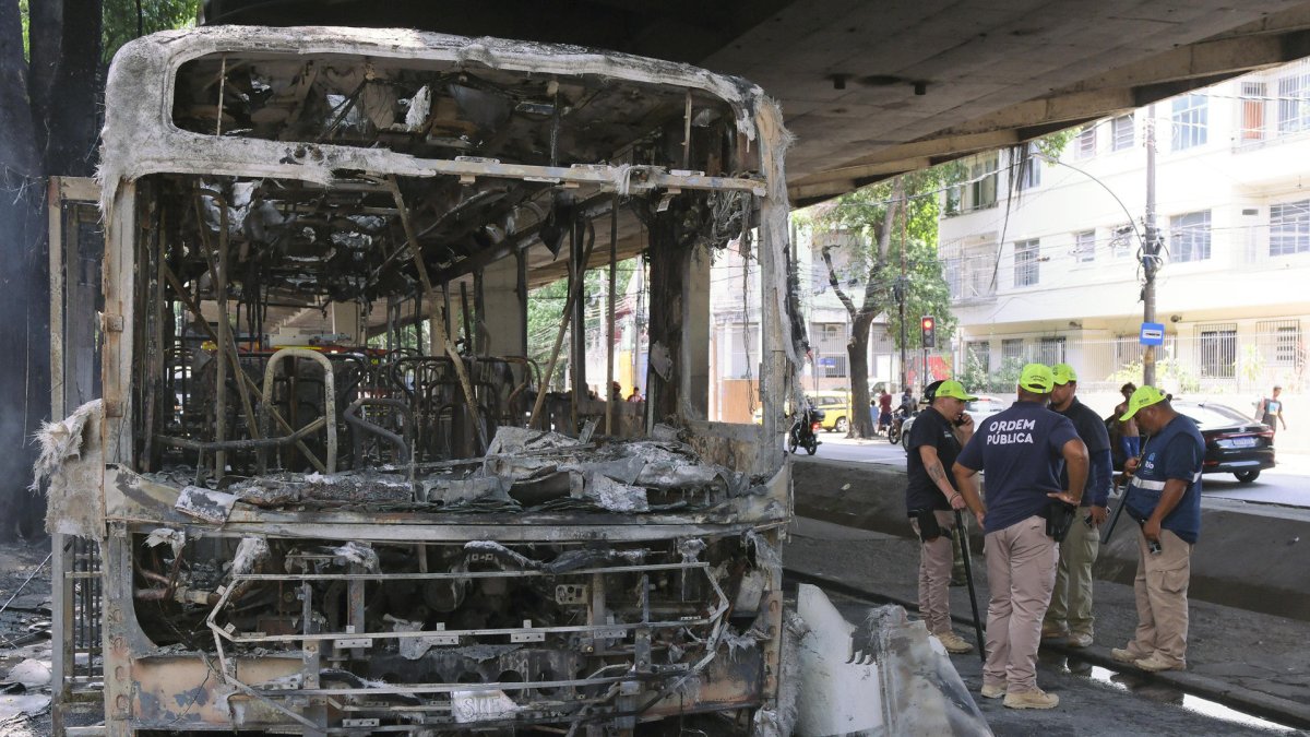 Bomberos observan los restos de un autobús incendiado por supuestos integrantes de una facción criminal, en respuesta a la muerte de Claudio Augusto dos Santos en Río de Janeiro.