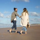 Side view of Caucasian couple on summer vacation. Bearded man and woman in casual clothes walking on beach, holding hands. Love, travelling, tenderness concept