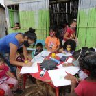 En una casa de caña, con pisos de tierra, 20 niños reciben ayuda educativa en la cooperativa Realidad de Dios. Líderes comunitarios colaboran en la tarea.