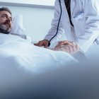 Hospitalized man lying in bed while doctor checking his pulse. Female physician examining male patient in hospital room.