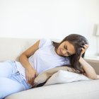 Woman in painful expression holding hands against belly suffering menstrual period pain,sitting sad on home bed, having tummy cramp in female health concept