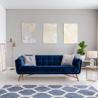 An elegant navy blue sofa in the middle of a bright living room interior with gold metal side tables and three paintings on a gray wall. Real photo.