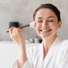 Smiling happy young woman applying makeup on her face with cosmetics brush in white spa bathrobe making morning preparations. Beauty care and treatment