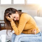 Young sick woman with hands holding pressing her crotch lower abdomen. Medical or gynecological problems, healthcare concept. Young woman suffering from abdominal pain while sitting on sofa at home