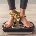 Cropped image of woman feet standing on weigh scales, on gray background. Legs winded with a tape measure