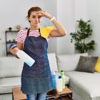 Young brunette woman wearing apron holding cleaning products at home worried and stressed about a problem with hand on forehead, nervous and anxious for crisis