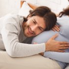 Pregnancy And Expectation concept. Future dad listening belly of his pregnant wife while lying on bed together, panorama