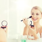 beauty, make up, cosmetics, morning and people concept - smiling young woman applying blush with makeup brush and looking to mirror at home bathroom