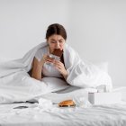 Sad hungry caucasian young lady eating chocolate bar on bed with fast food at home, suffering from stress, tension. Overeating, junk meal, depression and eating disorders. Health and mental problems