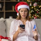 Worried concerned girl in Christmas Santa hat having problems with payment by credit card online for New Year purchases, looking at smartphone screen with puzzled face. Scam, fraud concept