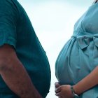 Hombre con sobrepeso y mujer embarazada, frente a frente, sosteniendo sus vientres con sus manos.
