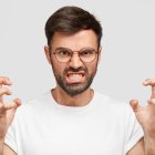 Portrait of emotive annoyed irritated unshaven male clenches teeth and gestures angrily while argues with wife, has irritated negative facial expression, wears casual white t shirt, stands indoor