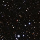 Imagen del protocúmulo Telaraña visto por la NIRCam (Near-InfraRed Camera) de Webb. Cientos de galaxias aparecen en esta vista, que se sitúa sobre el fondo negro del espacio. Hay muchos objetos superpuestos a distintas distancias. Incluyen grandes estrellas azules en primer plano, algunas con ocho picos de difracción, y galaxias espirales y elípticas blancas y rosas. En toda la escena aparecen numerosos puntos diminutos de color naranja]. Crédito: