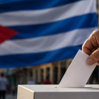 Una persona deposita su voto en una urna, con la bandera de Cuba de fondo, en una representación sobre el debate en torno al sistema electoral de la isla.