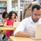 Diverse adult students working on computer in classroom. Focused sitting at desks and using laptops. Bookshelves in background. Education concept